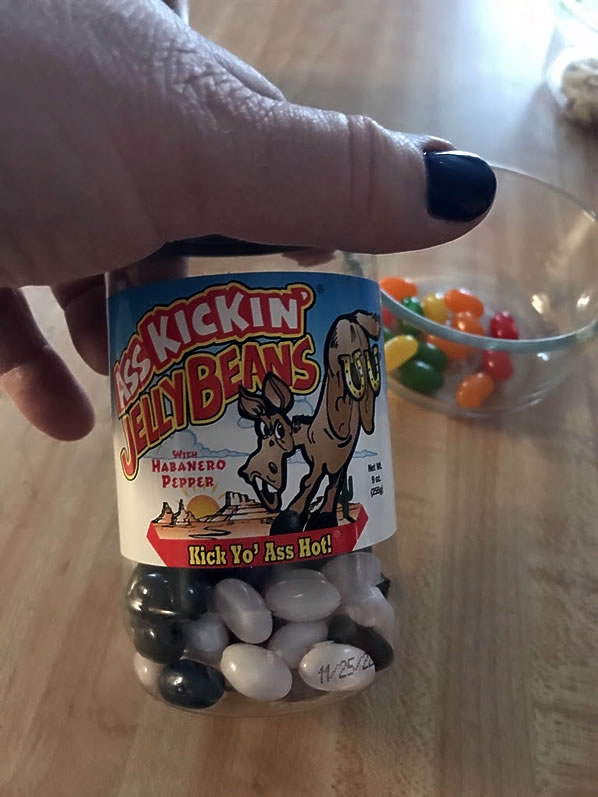 Penny's hand holding the jar of Ass Kickin' Jelly Beans with only black and white beans remaining in it; in the background is the bowl of fruit-flavored beans